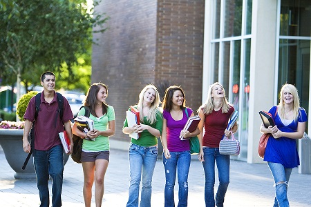 Six Students, one male and five females, walking out of school carrying their books talking and laughing. Horizontally framed photo.